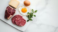 Fresh raw ribeye steaks with sunny side up egg, cheese block and herbs on white marble cutting board, top view composition for food photography and menu design.