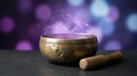 Tibetan singing bowl with wooden mallet releasing magical purple sparkles and glowing particles against blurred bokeh background for meditation and sound healing.