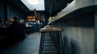 Empty bar stool at counter in modern industrial pub or restaurant interior with moody lighting and blurred background patrons socializing.