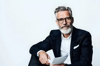Confident mature businessman with gray beard and stylish glasses holding digital tablet against white background, wearing black blazer and white shirt.