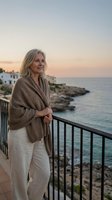 Mature blonde woman in cozy cardigan enjoying peaceful sunset moment on Mediterranean coastal balcony overlooking turquoise sea and rocky cliffs.