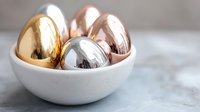 Metallic eggs in gold, silver, and rose gold colors nestled in white ceramic bowl on marble surface. Modern minimal Easter or luxury decor concept.