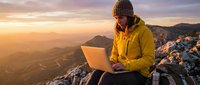 Remote worker in yellow jacket using laptop on mountain summit at sunrise, freelance professional working outdoors with scenic valley landscape.