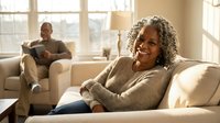 Smiling mature African American woman relaxing on sofa at home with husband reading in background, comfortable living room setting for lifestyle content.