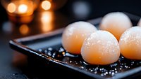 Delicate pink mochi dessert balls dusted with powdered sugar on dark ceramic plate with bokeh lights in background, elegant Japanese sweets presentation.