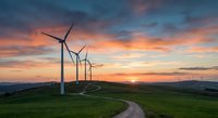 Wind turbines on green hills at sunset with dramatic orange and pink sky. Renewable energy landscape with winding road through countryside.