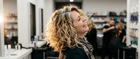 Young blonde woman with curly hair laughing joyfully in modern hair salon with stylists working in background. Professional beauty and wellness concept.