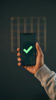 Hand holding smartphone displaying bright green checkmark on dark screen against blurred striped background. Represents approval, success, confirmation, and validation for app development, digital services, and technology concepts.