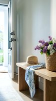 Cozy entryway with wooden bench, straw hat, floral scarf and lilac bouquet in wicker basket near glass door for home interior design.
