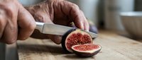 Hands cutting fresh fig in half with knife on wooden cutting board, close-up food preparation shot.