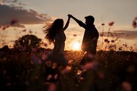 Romantic couple dancing at sunset in flower field, silhouetted against golden sky. Young man and woman enjoying intimate moment outdoors during golden hour.