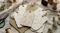 Handmade seed paper gift tags with natural twine, surrounded by crafting supplies including scissors, dried flowers, and wooden spools on linen fabric.