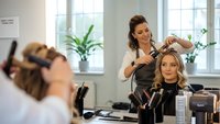 Professional hairstylist styling blonde woman's hair in modern salon with natural lighting and plants creating relaxing atmosphere for beauty services.