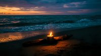 Cozy campfire burning on sandy beach at sunset with dramatic purple and orange sky reflecting on ocean waves creating romantic evening atmosphere.