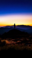 Silhouetted figure standing on mountain peak at sunrise, gazing over layered mountain ranges with golden and blue gradient sky, ideal for motivational and travel concepts.
