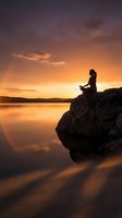 Woman sitting on rocky outcrop meditating at golden sunset over calm lake, silhouetted against orange sky with mountains in distance. Wellness and mindfulness concept.
