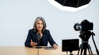 Mature woman with gray hair and South Asian ethnicity, wearing a dark blazer, speaking at a desk with a microphone during a professional video recording or broadcast setup with camera equipment.