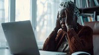 Young African American woman experiencing stress while working from home, holding head in hands near laptop, wearing cozy sweater in moody natural lighting.