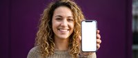 Young Caucasian woman with curly hair smiling and showing smartphone with blank white screen on purple background.