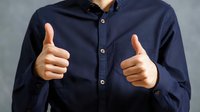 Person in navy blue button-up shirt showing thumbs up with both hands against gray background, conveying positive approval or agreement.