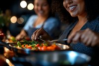 Young African American woman smiling while preparing fresh vegetables for dinner with friend. Warm evening atmosphere with bokeh lights creates cozy dining experience.