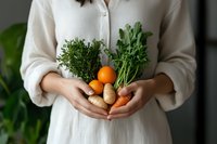 Young Asian woman in white linen dress holding fresh organic vegetables and herbs from garden harvest including oranges, potatoes, carrots and green herbs.