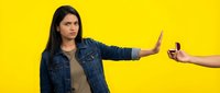 Young Hispanic woman in denim jacket making stop gesture refusing smartphone on bright yellow background. Concept of digital detox and boundaries.