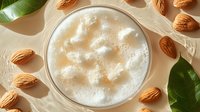 Frothy almond milk latte in glass cup from above, surrounded by scattered raw almonds and fresh green leaves on beige background, natural light.