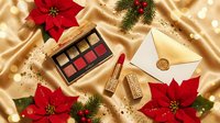 Luxury Christmas makeup collection with red lipstick, eyeshadow palette and golden envelope on silk fabric with poinsettia flowers and pine branches.