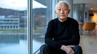 Senior Asian man in black sweater sitting thoughtfully by large window in modern interior, arms crossed, natural lighting creates contemplative atmosphere.