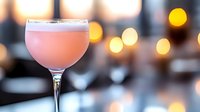 Pink cocktail in elegant coupe glass with sugar rim on bar counter, warm bokeh lights creating sophisticated evening atmosphere for mixology and nightlife.