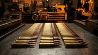 Rows of golden brass ammunition cartridges arranged on metal surface in industrial manufacturing facility with machinery in background.