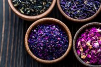 Dried purple and blue flower petals and tea leaves in wooden bowls on dark wooden background, top view composition for wellness and aromatherapy concepts.