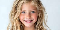 Cheerful young child with wavy blonde hair and bright blue eyes smiling against white background, conveying happiness and innocence.