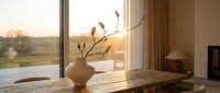 Warm sunlight streams through modern windows onto wooden dining table with ceramic vase and dried branches in contemporary home interior during golden hour.