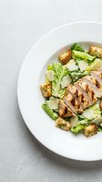 Grilled chicken breast sliced and served over fresh romaine lettuce with croutons and parmesan cheese on white plate, overhead view for restaurant menu or food blog.