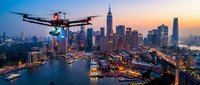 Drone flying over illuminated city skyline at sunset with skyscrapers reflecting in water. Modern aerial technology for urban photography and surveillance applications.