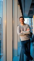 Confident young Caucasian businessman in casual sweater standing with crossed arms in modern office with city skyline view through floor-to-ceiling windows.