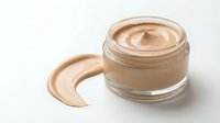 Open glass jar of beige foundation cream with smooth swirl texture and makeup smear on white background for cosmetic product advertising.