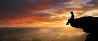 Silhouetted figure sitting on mountain cliff edge at dramatic sunset with golden sky and misty valleys below, conveying freedom and contemplation for motivational and adventure content.