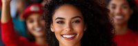 Young African American woman with curly hair smiling at camera in crowd of diverse people wearing red clothing at sports event or rally gathering.
