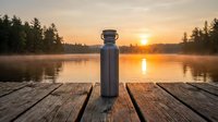Insulated water bottle on wooden dock at sunrise over misty lake surrounded by evergreen forest, ideal for outdoor lifestyle and beverage product marketing.