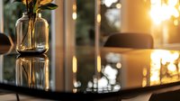Glass vase with green leaves on reflective surface at sunset, creating warm atmospheric mood with bokeh lights and window reflections in modern interior setting.