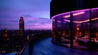 Luxurious rooftop restaurant with panoramic city views at twilight, illuminated by purple and pink neon lights against dramatic sunset sky and glowing urban skyline.