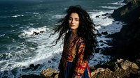 Young woman with long curly dark hair wearing traditional ethnic patterned clothing standing on rocky coastline with ocean waves crashing below.