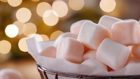 Fluffy white marshmallows in metal basket with soft golden bokeh lights creating warm cozy atmosphere, macro food photography for winter treats.