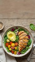 Grilled chicken breast sliced over mixed greens, avocado, tomatoes, and red onion in a ceramic bowl on wooden surface for healthy lunch meal prep.