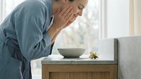 Mature woman with light skin tone, wearing casual blue shirt, touching face with hand in gesture of concern or discomfort while looking at empty bowl on wooden dresser near window. Suitable for healthcare, wellness, and lifestyle editorial use.