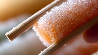 Macro shot of crystallized sugar coating on salmon sashimi held with metal chopsticks, showing delicate texture and culinary detail in soft focus background.