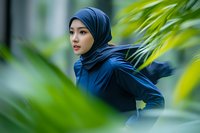 Young Asian woman in navy blue hijab and athletic wear looking determined while exercising outdoors among lush green tropical plants in soft natural lighting.